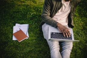 Man on grass with laptop on lap and notebooks beside him. Ethically Market Your Therapy Practice.